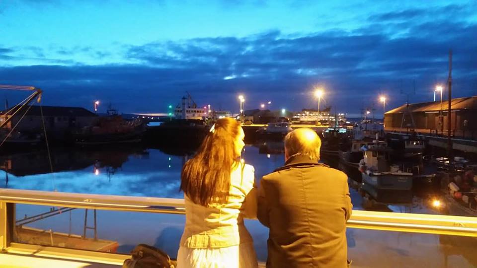 a man and woman looking out over Kirkwall Harbour at night with the lights of the vessels
