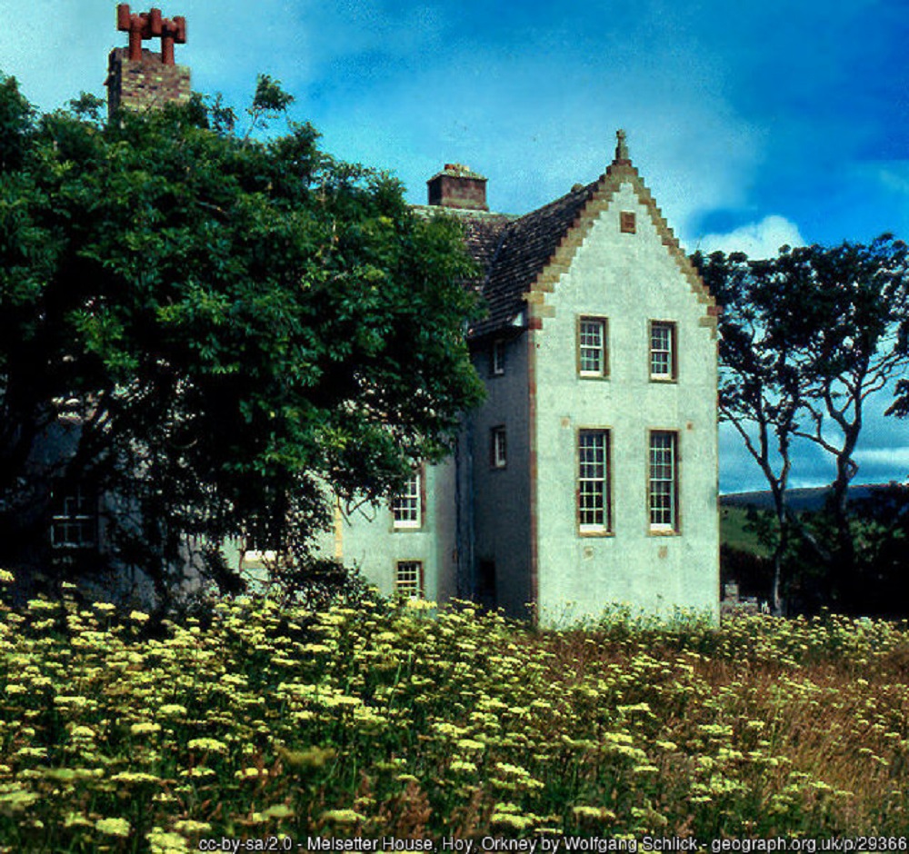 The south gable of Melsetter House Hoy surrounded by yellow flowers and trees to the right. The gable end has four sets of windows, 2 larger ones on the ground floor and 2 smaller above