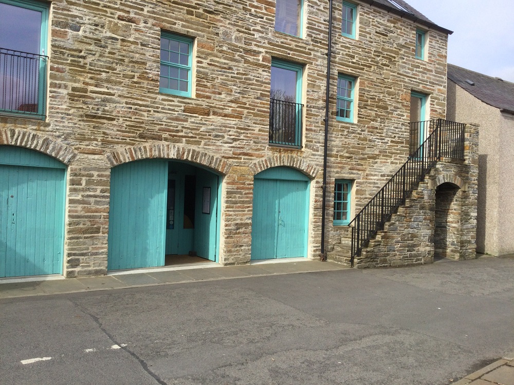The renovated listed warehouse building showing the stone exterior, arched doorways with green painted wooden doors. a staircase leads up to a door. windows above some with railings.