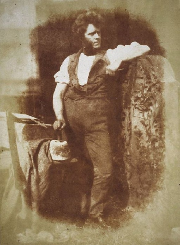 Photograph of Hugh Miller, he stands with one arm resting on a huge upright stone and another with his hand resting on a large stone weight he is dressed in working clothes with his sleeves rolled up