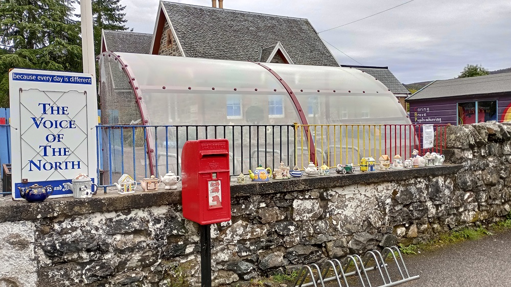 A post box on a stand next to a wall covered in small tea pots next to the local school and a polytunnel