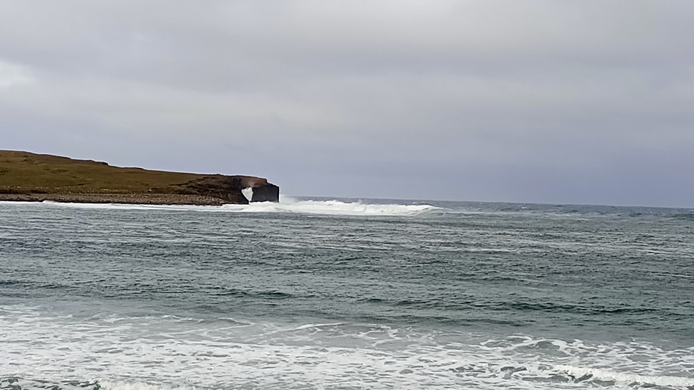 the bay of Skaill with stormy seas and cloudy skies as waves crash through the Hole o Roe in the distance