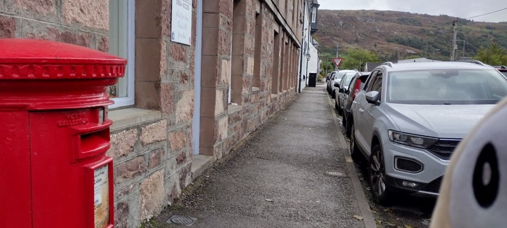 a pillar box on the side of the pavement looking up the street in Ullapool with the hills in the background