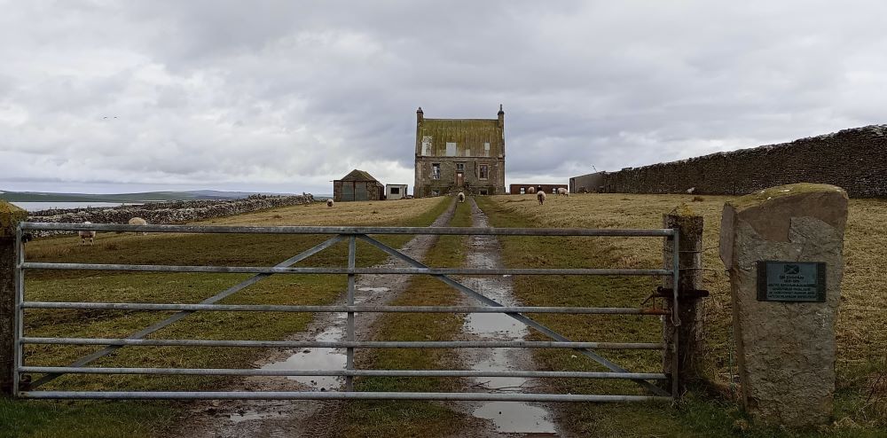The large house from the end of the path with its asbestos covered roof and muddy tracks. sheep graze on the grass