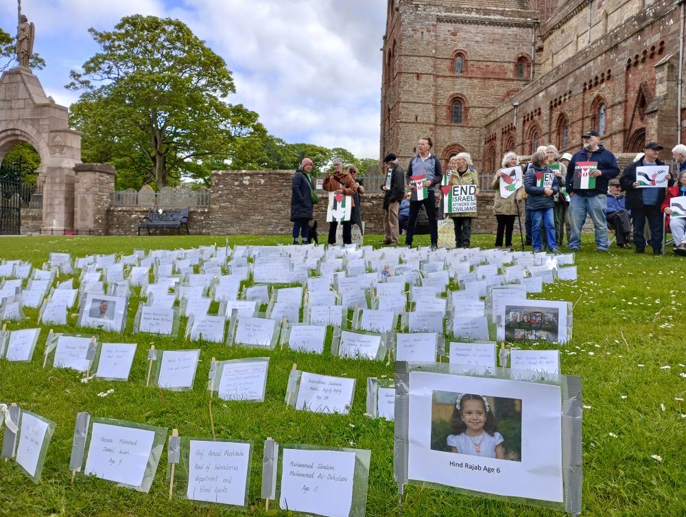 the names of many killed are to the foreground as the image looks up towards those in vigil outside the Cathedral