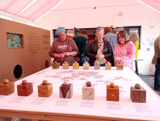 three women look at the display of layered cubes and balls of clay in front of a geological map