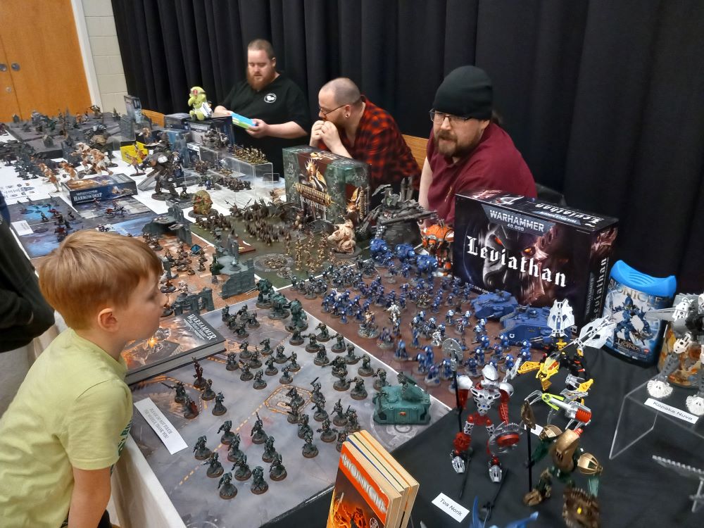a young boy looking at a large display of Warhammer models and three exhibitors behind the display with one speaking about the models