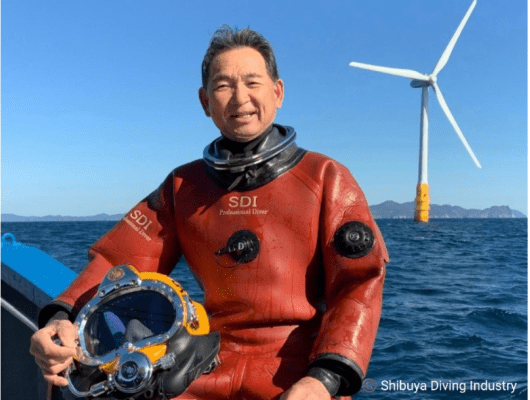 Masanobu Shibuya in diving gear on a boat with a wind turbine behind him