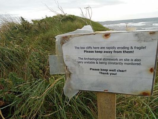 sign on a grass covered dune. The low cliffs here are rapidly eroding and fragile. Please keep away from them.