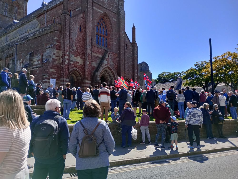 crowds of people in front of the St Magnus Cathedral where people stand on the steps with the flags of Norway, Orkney and Scotland