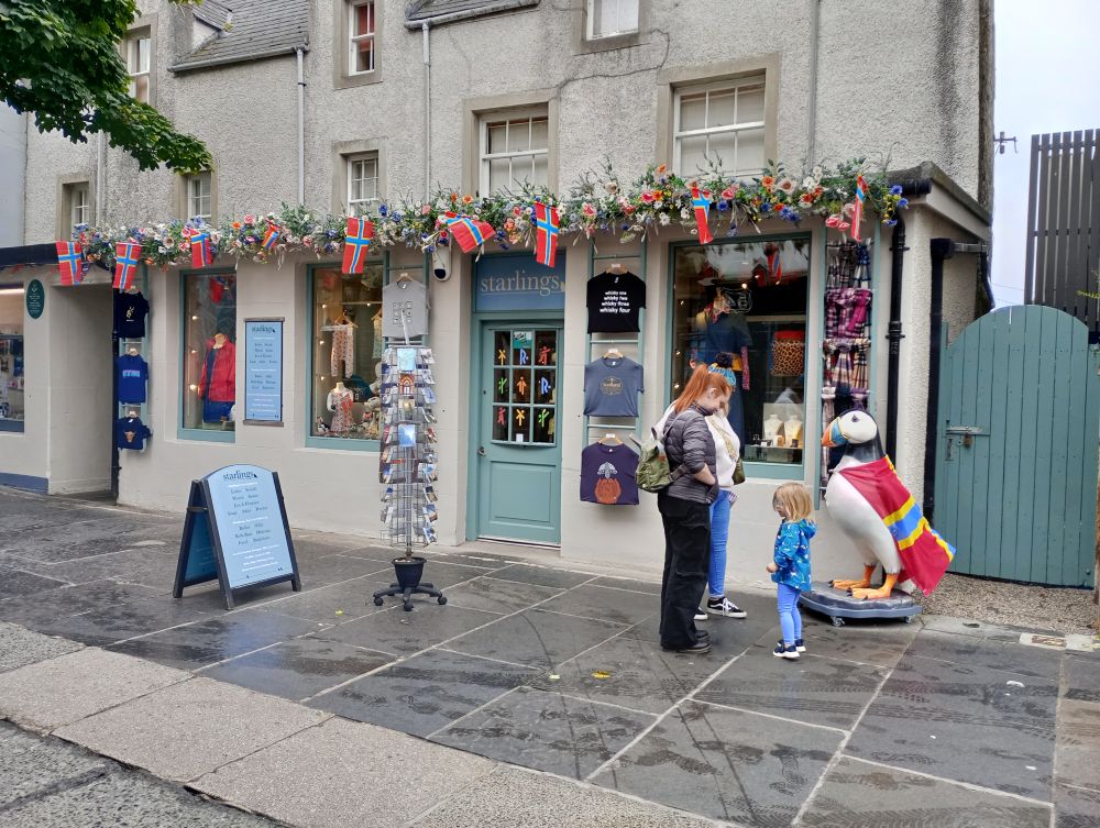 two women and a small child standing outside the Starlings shop beside a large stuffed puffin wearing an Orkney flag