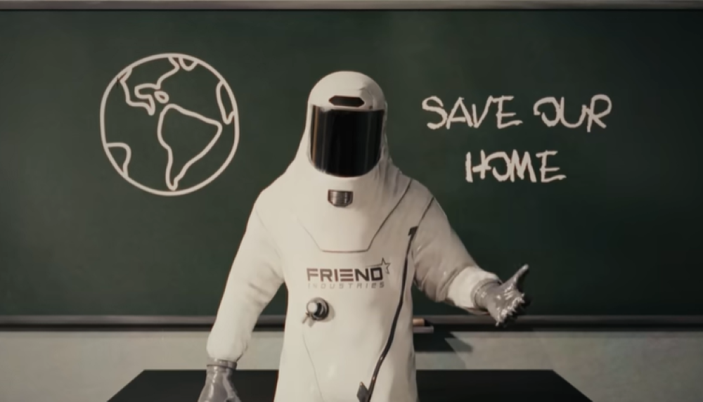 screen shot from The Moon Woke Up with an astronaut fully suited with Friend on his suit standing in front of a blackboard that says save my home and a drawing of Earth