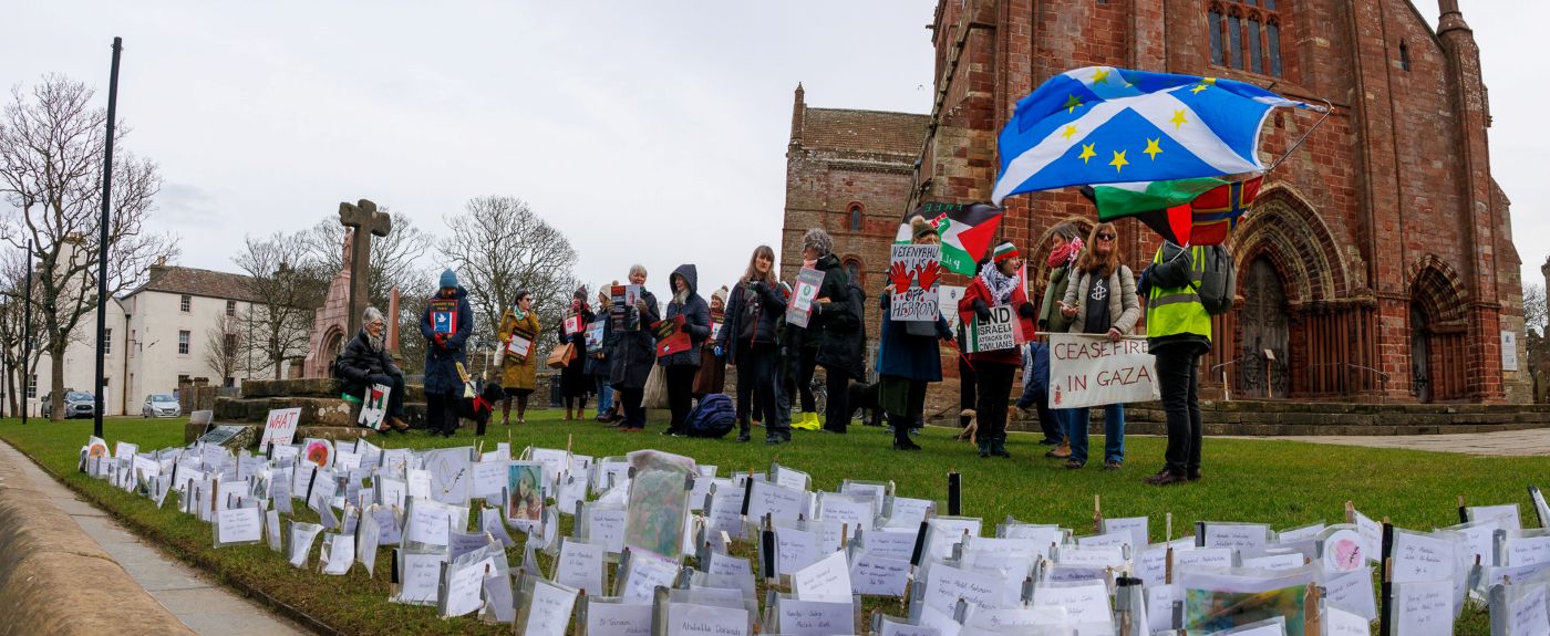 the names of thsoe killed and behind them people standing in vigil with posters and flags